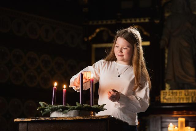 Jente med lys genser står i en relativt mørk kirke og tenner lys i en adventskrans.